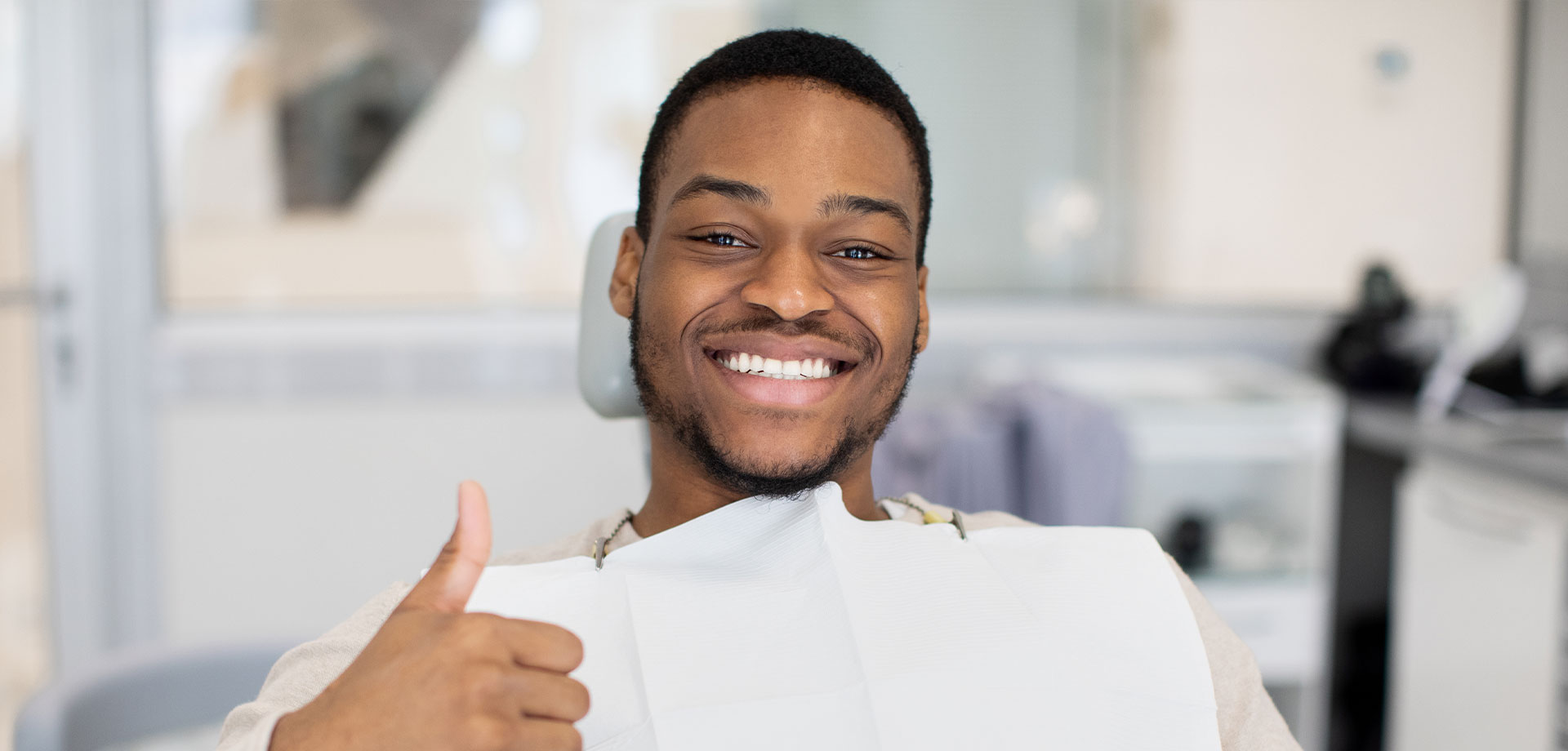 The image shows a man sitting in a dental chair, giving a thumbs-up gesture with a smile on his face.