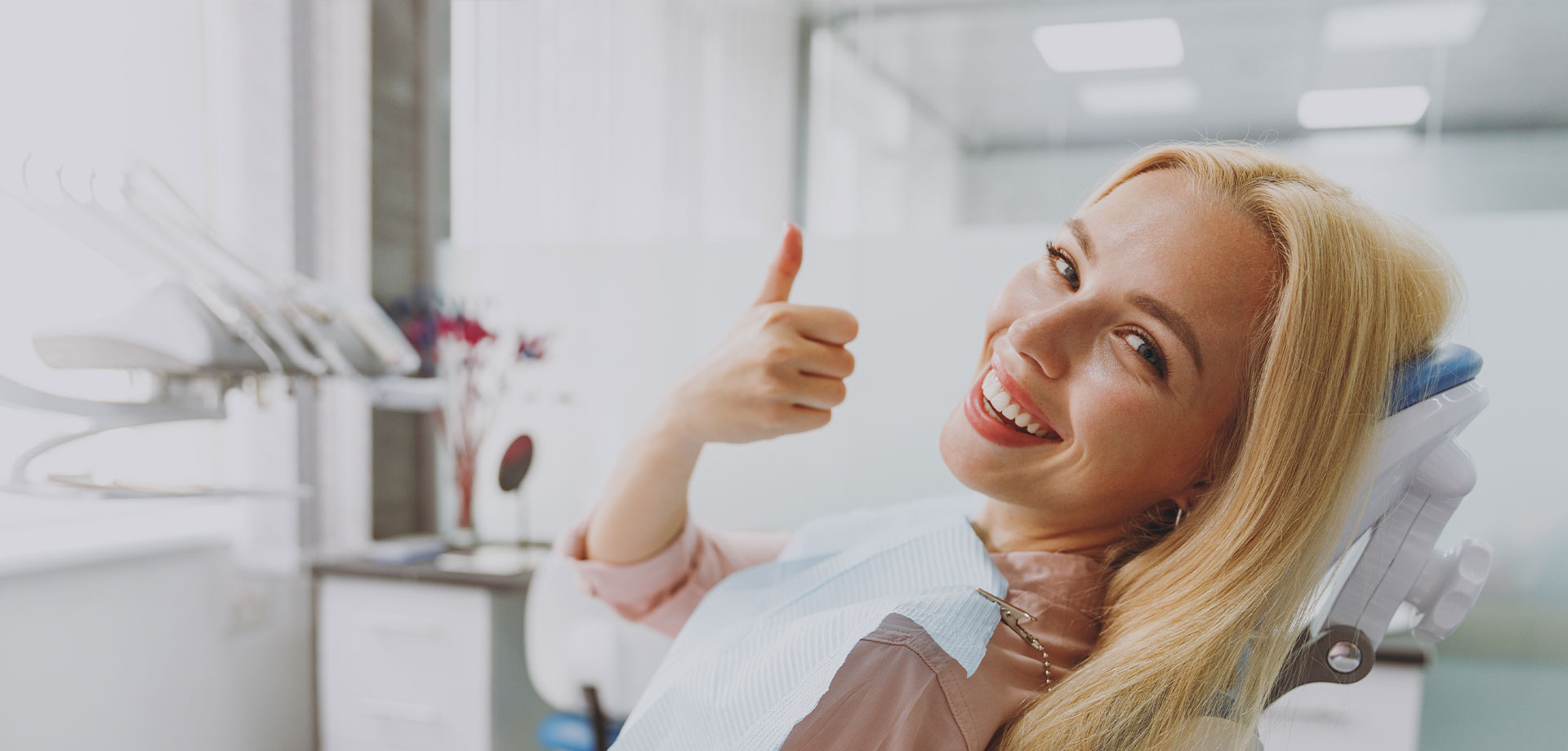 A smiling woman in a dental office, giving a thumbs-up sign.