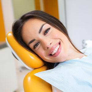 A woman with a broad smile is sitting in an orthodontist s chair, wearing a blue surgical mask and headgear.