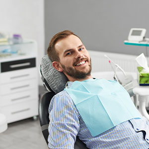 A man is sitting in a dental chair with a smiling expression, wearing a blue surgical mask and holding a mouth mirror.