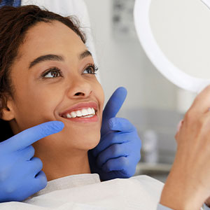 The image depicts a young woman with a radiant smile, sitting in a dental chair and receiving dental care from a professional.