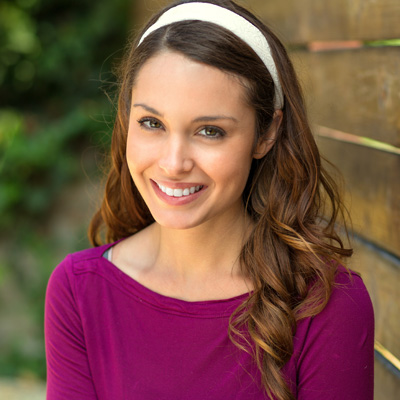 A young woman with long brown hair, wearing a purple top and a white headband, smiling at the camera.