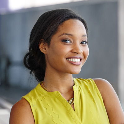 The image shows a woman smiling at the camera, wearing a yellow top and standing in front of a gray wall.