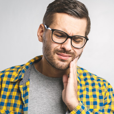 Man with beard and glasses, wearing a plaid shirt, holding his hand to his mouth in a gesture of concern or pain.