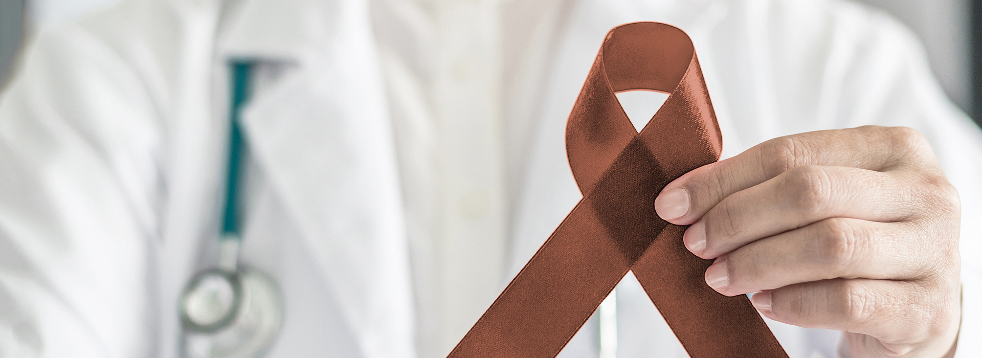 The image shows a person in a white coat holding a red ribbon with a brown bow, which is commonly associated with awareness for cancer.