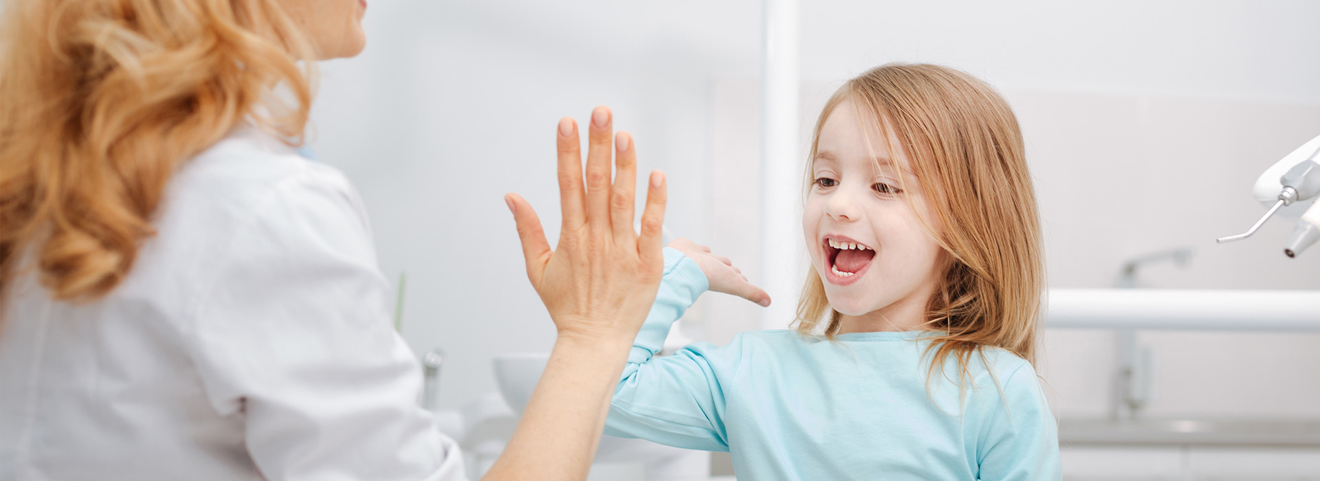 The image is a photograph of a woman and a young girl in an indoor setting, likely a dental or medical office, with the woman wearing a white coat and the girl looking at her with a smile.