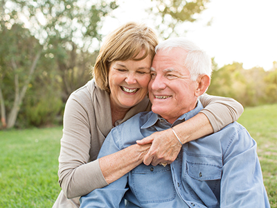The image shows an elderly couple embracing each other outdoors, with the man wearing a blue shirt and the woman in a beige top.