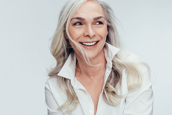 The image features a woman with short hair, smiling at the camera. She has grey hair and is wearing a white top.