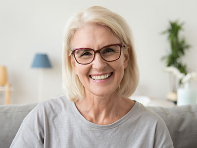 The image features a smiling woman with short blonde hair, wearing glasses and a light-colored top, seated indoors against a white wall.