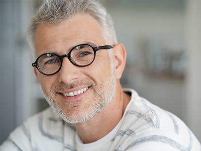 The image shows a man with glasses, smiling slightly at the camera. He has short hair and is wearing a white shirt with a patterned collar.