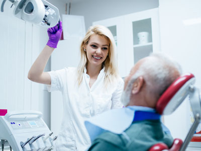 In the image, a woman in a white lab coat is standing next to an elderly man who is seated in a dental chair. She appears to be a dentist or dental assistant, holding up a dental mirror and examining his mouth. The setting suggests this could be a dental office.