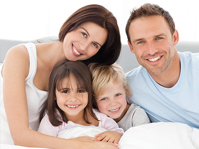 A family of four, including two adults and two children, smiling together in a bedroom with a white bedspread.