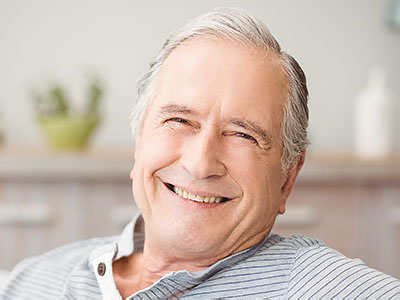 The image shows an older man with white hair, smiling and looking directly at the camera. He is wearing a blue shirt and appears to be sitting in a relaxed posture.