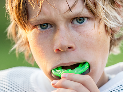 This is a photograph of a young boy with blonde hair, wearing a football jersey and holding a green sports mouthguard in his mouth.