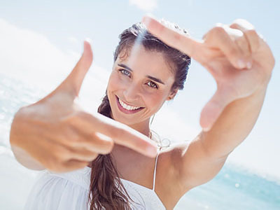 A woman is standing on a beach, holding her hand up to the camera with her fingers forming a circle.